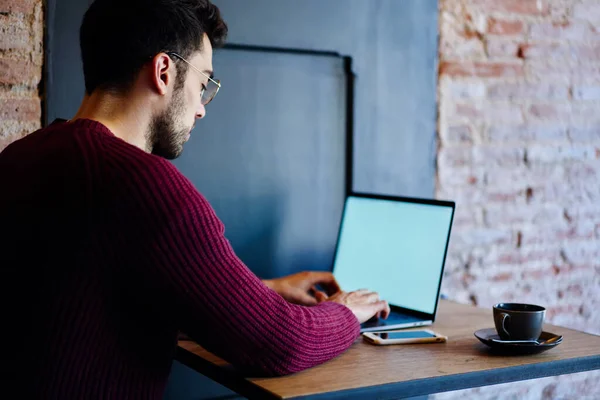 穿着紫色毛衣和眼镜 头脑清醒的年轻男性员工在明亮舒适的咖啡店里工作时 用空白屏幕在笔记本电脑上打字 — 图库照片