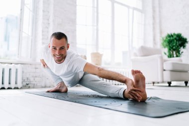 Neşeli beyaz erkek kameraya bakıyor kaslarını esnetiyor ve vücudunu evde çalıştırıyor. Dairesinde gülümseyen genç hippi, sağlıklı bir zihin ve beden için Asna 'da duruyor.