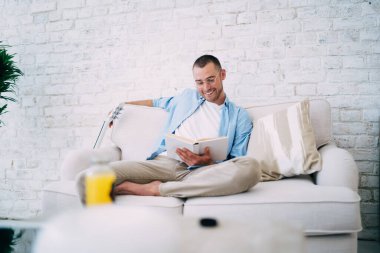 Keyifli bir erkek, rahat bir kanepede dinlenirken gülümseyerek ve ilginç kitaplar okuyarak.