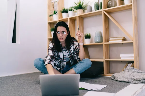 Serious female remote employee sitting cross legged with pen in hand and solving problem via laptop while working at home