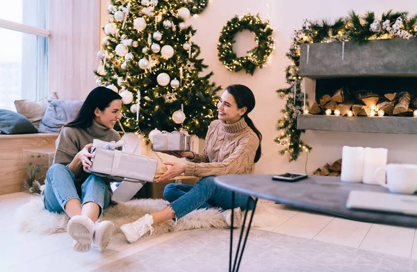 Excited ladies sitting in decorated living room near fireplace with wrapped gift boxes and celebrating Christmas holiday while looking at each other