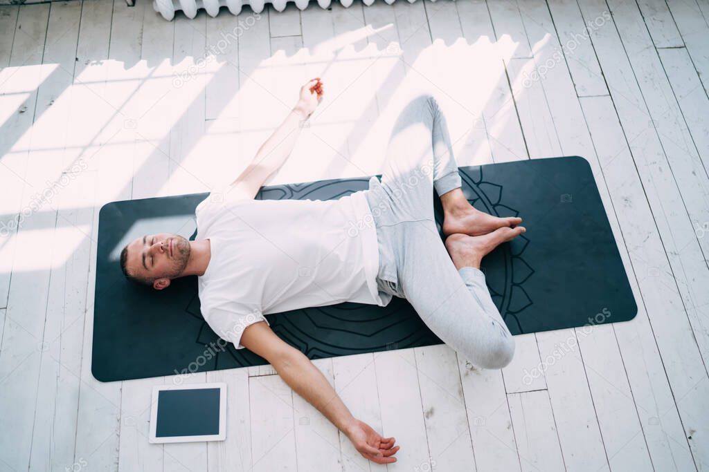 From above of serene male yoga guru lying on rubber mat near tablet with empty screen and performing Supta Baddha Konasana pose
