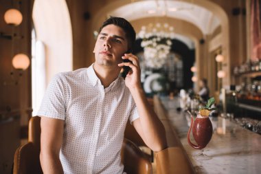 Gündüz vakti bar tezgahının yanındaki kafede kokteyl içerken akıllı telefondan konuşan, gündelik kıyafetli, kendine güvenen bir adam.