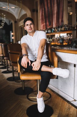 Rahat genç erkek rahat kıyafetler ve spor ayakkabılar içinde şık bir kafede bacak bacak bacak üstüne atmış bir şekilde otururken başka tarafa bakıyor.