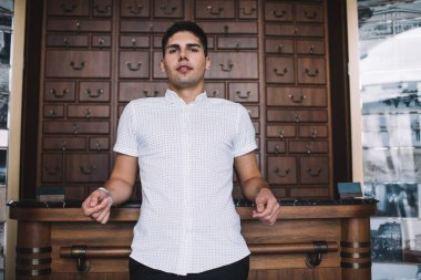 Kendine güveni tam bir adam ahşap bir dolaba yaslanmış, içinde retro bir iç mekan olan ve randevu beklerken kameraya bakan.