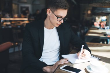 Odaklanmış koyu renk saçlı erkek resmi kıyafetler ve gözlüklerle kafede elinde kalemle otururken deneme üzerinde çalışıyor.