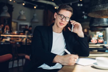 Kendine güveni tam bir adam, modern bir restoranın masasında oturmuş, iş projesinde not defterine planlar yazıyor ve cep telefonuyla konuşuyor.