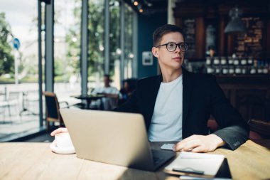Siyah ceketli ve gözlüklü, kafeteryada kahve içip gözlerini kaçıran ciddi bir erkek işçi.