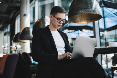 İyi giyimli gözlüklü öğrencinin elinde sıcak içecekle masada otururken dizüstü bilgisayarla çalışmasından.