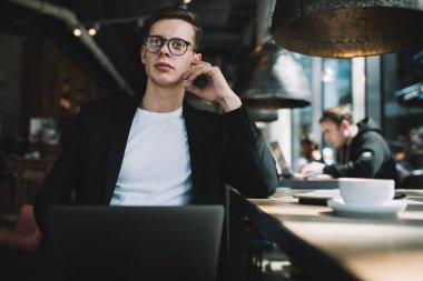 Ciddi, gözlüklü, dizüstü bilgisayarlı genç bir adam ara sırasında modern kafede bir fincan kahve içerken kameraya bakıyor ve düşünüyor.