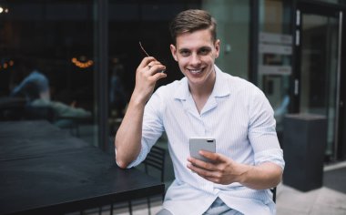 Beyaz gömlekli yakışıklı genç adam akıllı telefon kullanıyor ve modern kafenin terasında cam duvarlı kameraya bakıyor.