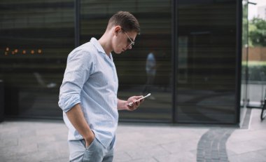 Gündelik kıyafetli ve güneş gözlüklü bir erkeğin cep telefonuyla internette gezinirken kaldırımda el ele tutuşup güneş ışığının altında duruşu.