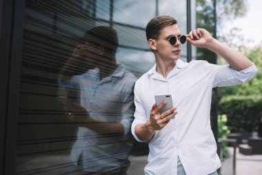 Tişörtlü, cep telefonlu, güneş gözlüklü genç adam öğleden sonra karanlık, şeffaf bir duvara yaslanmış ve gözünü kaçırmıştı.