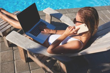 Havuz başında güneşli bir günde havuz başında dinlenirken, cep telefonuyla konuşan ve sörf yapan, sarı saçlı, mayo ve güneş gözlüklü, tanınmamış bir kadından.