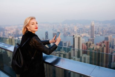 Hong Kong şehir merkezindeki çağdaş binanın çatısında sırt çantalı ve cep telefonuyla fotoğraf çekerken yüzünü çeviren içerikli kadın turistin yan görüntüsü