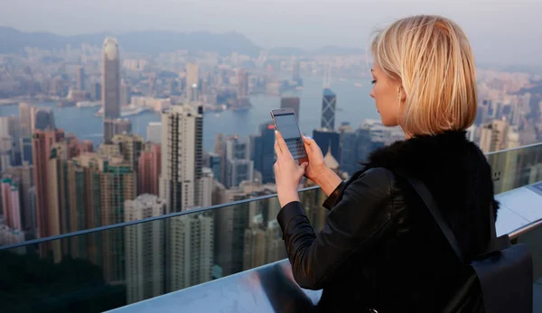 香港のダウンタウンにある現代的な建物の屋上に立ち 携帯電話でネットワークを閲覧するバックパックとスタイリッシュな服で女性旅行者のバックパックビュー — ストック写真