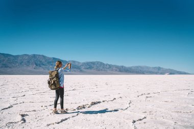Sırt çantasıyla Badwater Havzası 'nın çorak zemininde duran ve sıcak bir günde sonsuz manzaranın fotoğrafını çeken tanınmamış bir kadın yürüyüşçünün arka görüntüsü.