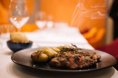 Izgarada pişirilmiş nefis sıcak et biberiyeyle süslenmiş fırında patatesle birlikte beyaz masa örtüsüyle kaplı siyah seramik tabakta servis ediliyor.
