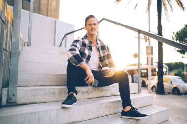 Günbatımında şehir caddesinde merdivenlerde oturup kahve içerken kameraya bakarak ve kulaklıkla kulaklık takarak müzik dinleyerek tatmin olmuş genç bir erkek.