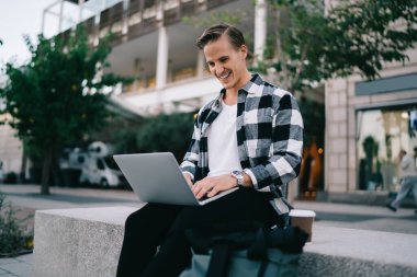 Neşeli blog yazarı sosyal ağlarda komik bir fotoğraftan gülüyor dizüstü bilgisayarda web sayfasına bakıyor, 20 'li yaşlarda gülümseyen beyaz hippi şehirde dizüstü bilgisayardan eğitim kursu öğreniyor.