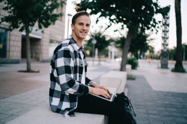 Sokakta bankta oturan ve iş sırasında dizüstü bilgisayara bakıp günlük kıyafetlerle kameraya bakan genç adamın yan görüntüsü.