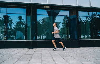 Kentteki kardiyo eğitimi sırasında koşan kararlı erkek koşucunun yandan görünüşü, fiziksel yoğunluk üzerinde yoğunlaşmak için ısınma sırasında dayanıklılık egzersizi yapan beyaz bir adam.