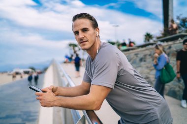 Gülümseyen 20 'li yaşlardaki yakışıklı hipster adamın spor molasında dinlenen portresi. Uygulamadan müzik dinlemek için cep telefonu kullanıyor. Pozitif sportmen açık havada akıllı telefondan koşmak hakkında podcast yapmaktan hoşlanıyor.