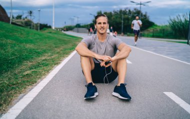 Yakışıklı koşucunun portresi asfalt yolda koşmak ve kameraya bakmak için dinleniyor, elektronik kulaklıklı kaslı sporcu ses eğitimi almış müzik podcast 'i dinliyor ve poz veriyor.