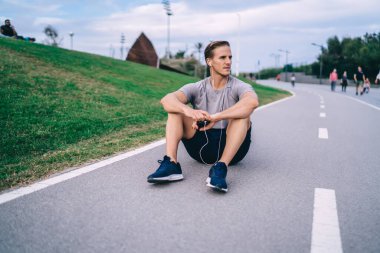 Elektronik kulaklıklı beyaz bir adam asfalt yolda dinleniyor. Kardiyo eğitimi için ses tavsiyesi dinliyor. 30 'lu yaşlarda kulaklıklı bir erkek antrenman sırasında medya uygulaması kullanıyor.