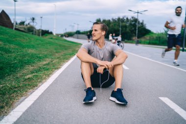 Elektronik kulaklıklı beyaz bir adam asfalt yolda dinleniyor. Kardiyo eğitimi için ses tavsiyesi dinliyor. 30 'lu yaşlarda kulaklıklı bir erkek antrenman sırasında medya uygulaması kullanıyor.