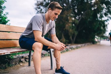 Kulaklıklı beyaz bir erkek, sokak bankında dinlenen müzik şarkılarını seçiyor, kulaklıklı bir sporcu, modern cep telefonuyla kişisel antrenörle mesajlaşırken ses tavsiyesi dinliyor.