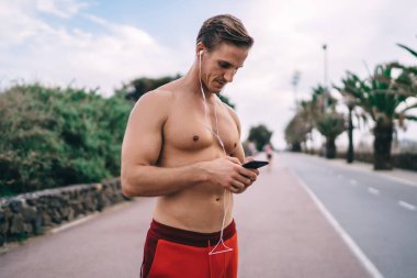 Seksi gömleksiz kaslı beyaz vücutçu motivasyon müzik listesi için hücresel teknoloji kullanıyor ve sabah antrenmanı sırasında kulak misafiri oluyor. Mobil uygulama kullanan kulaklıklı atletik adam.