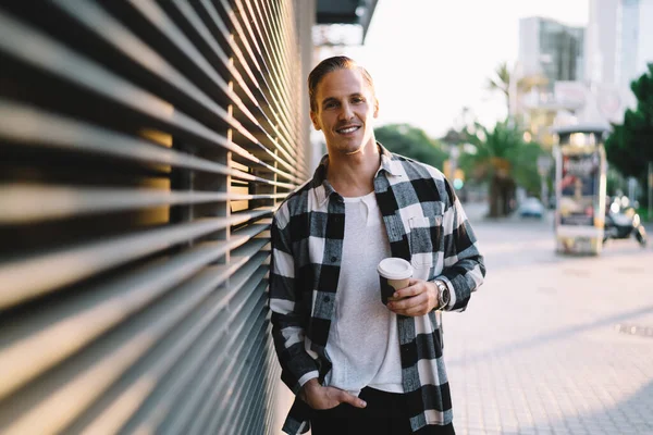Half length portrait of cheerful caucasian male dressed in casual trendy outfit holding coffee cup enjoying leisure in city,handsome smiling hipster guy looking at camera standing on urban settings