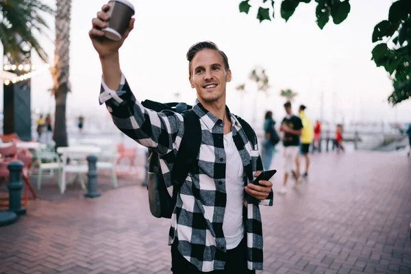 Happy caucasian hipster guy in trendy wear raising coffee cup offering beverage standing on street, smiling male traveler in casual outfit gesture and smiling exploring city on vacations on square