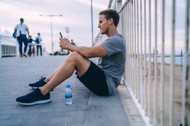 Genç hippi dışarıda koştuktan sonra dinleniyor. Sosyal ağlarda blog yazmak için akıllı telefonu kullanıyor. Dalgın erkek koşucu akıllı telefondan video izliyor.