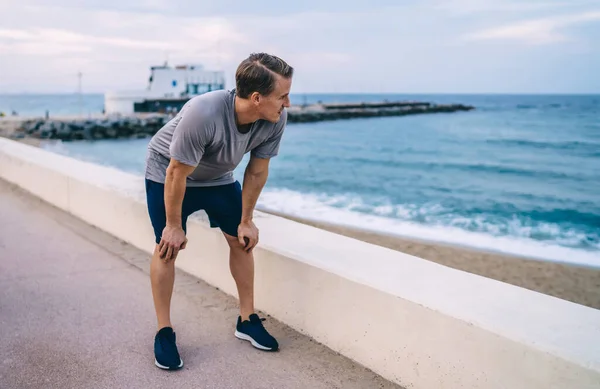 Pensive caucasian male jogger in activewear breathing after cardio workout looking at water in harbor, young slim sportsman breathin and refreshing after running making pause break on quay in morning