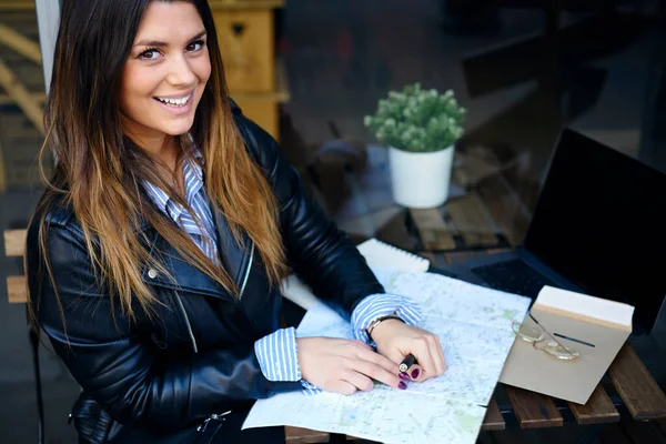 From above of happy young female with brown hair in casual wear smiling and sitting at table with city map