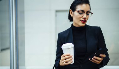 Zarif siyah ceketli ve gözlüklü konsantre genç bayan New York 'ta telefon ve kahveyle tek başına duruyor.