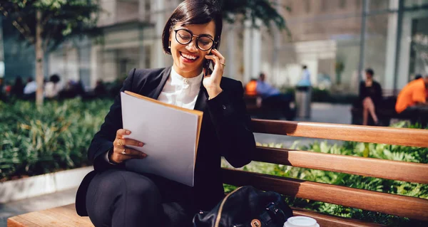 Laughing young woman in business suit and eyeglasses with cup of takeaway coffee sitting on bench and talking on smartphone while working with documents