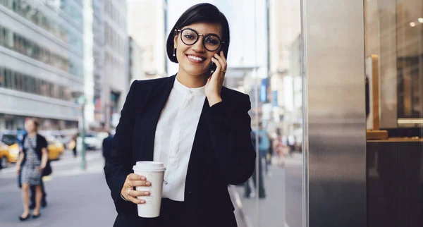 穿着正装和眼镜站在街上 一边用智能手机聊天一边喝热腾腾的外卖咖啡的积极时尚女性 — 图库照片