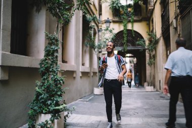 Moda kıyafetleri ve gözlükleriyle gülümseyen ve kulaklıklı kulaklıkla müzik dinleyen mutlu bir etnik erkek.