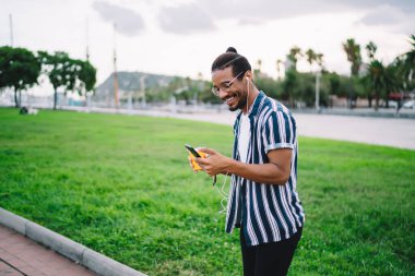 Kahveli mutlu koyu tenli erkek milenyum çocuğu cep telefonunda yeni şarkılar arıyor. Afro-Amerikan hipster aksesuar ile tatmin olmuş.
