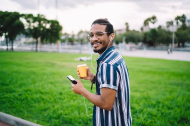 Neşeli genç etnik adam tek kullanımlık kahveyle parkta yürürken kulaklıklı cep telefonuyla müzik dinliyor.