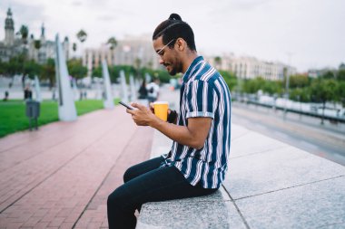 Pozitif esmer tenli erkek gezgin sokakta oturmuş boş zamanlarında gelen mesajları kontrol ediyor, gülümseyen Afro-Amerikan hipster adam akıllı telefon uygulamasından uyarı indiriyor.