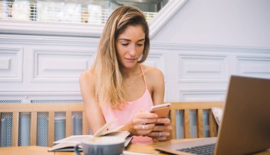 Modern kafede masada otururken ve kahve molası sırasında dizüstü bilgisayarda çalışırken gündelik kıyafetlerle gezen akıllı telefonlu bir kadın.