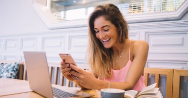 Neşeli gülümseyen etnik kadın blog yazarı gündelik kıyafetlerle dizüstü bilgisayarla proje üzerinde çalışıyor ve kafede otururken akıllı telefonla mesajlaşıyor.