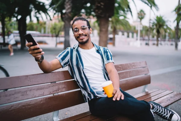 Cheerful handsome hipster guy in casual wear sitting on city bench resting with beverage and using smartphone, positive african american man looking at camera sending messages and mails via 4G