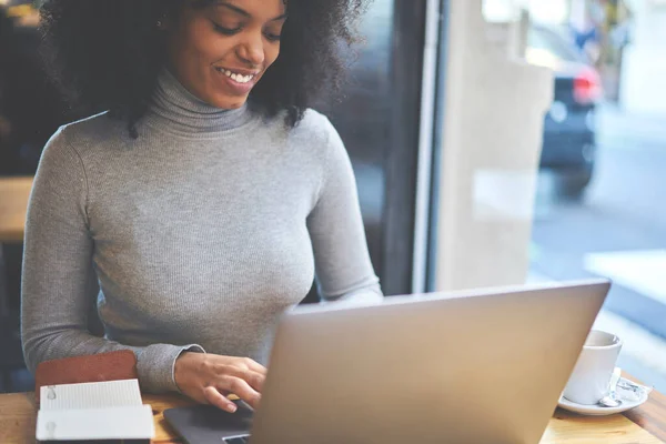 穿着休闲装的黑人女自由撰稿人坐在现代工作场所 一边带着上网本 一边笑着 一边在下班后上网交流 — 图库照片