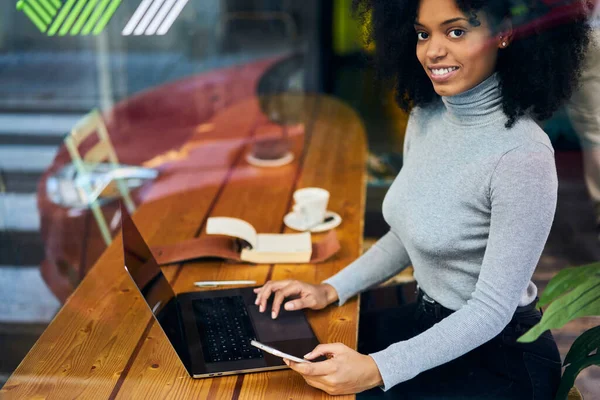 クロップ若いアフリカ系アメリカ人女性のフリーランスは ノートパソコンでリモートで作業し カメラを見ている間 居心地の良いワークスペースやブラウジングスマートフォンのテーブルに座ってカジュアルな服装で — ストック写真