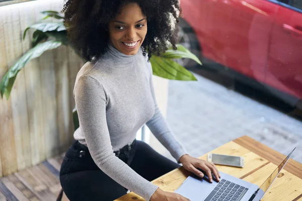 カジュアルな服を着たアフリカ系アメリカ人の女性のフリーランスの上から笑顔と離れてカフェでノートパソコンを入力しながら — ストック写真